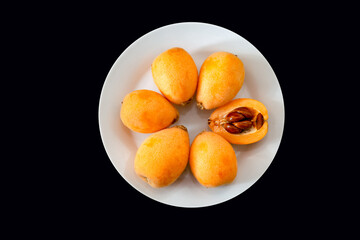 Ripe loquats on white plate on black background top view. One cut in half showing the inside of the fruit with the seeds ( Eriobotrya japonica )