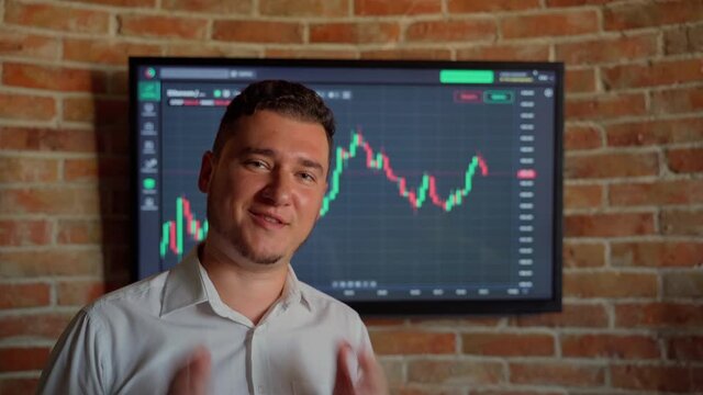 Man trader talk looking at camera graph finance background indoors. Male person has video call conference talking about currency online money. Quotes eth and btc at stock exchange market chart 