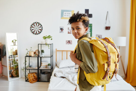 Waist up portrait of mixed-race teenage boy holding backpack and smiling at camera while getting ready for school, copy space