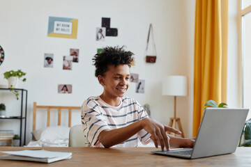 Side view portrait of mixed-race teenage boy using laptop and smiling in bright room interior, copy space © Seventyfour