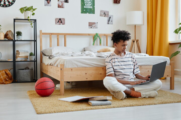 Full length portrait of mixed-race teenage boy sitting on floor in cozy room and using laptop, copy...