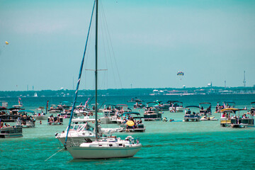 Variety of boats including sailboats parked, anchored off reef for boating party with turquoise water in Destin, Florida
