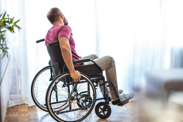 Fototapeta na wymiar Side view of mature man sitting in wheelchair and looking away. Side view of thoughtful disabled active Caucasian man looking through window on wheelchair in a comfortable home