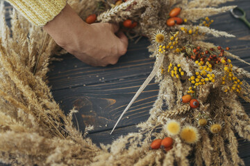 Hands making stylish autumn rustic wreath with dry grass, wildflowers, wheat and berries on rustic...