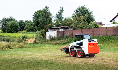 A skid steer loader clears the site for construction. Land work by the territory improvement. Machine for work in confined areas. Small tractor with a bucket for moving soil and bulk materials.