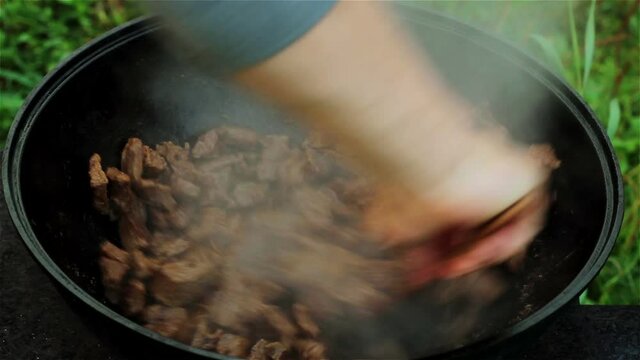 meat is stewed in a large cast-iron cauldron over an open fire. The beef is mixed with a wooden spoon and onions are added to the pan. All ingredients are mixed in the pot.