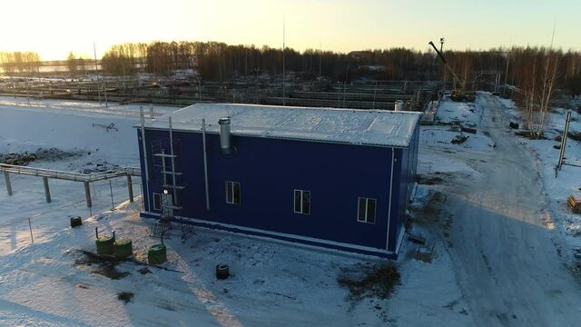 Blue workshop building with snowy roof chimney and pipelines at wastewater treatment plant at sunset in winter aerial view