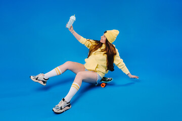 Fototapeta na wymiar a pregnant girl in yellow clothes with a glass of juice sits on a skateboard on a blue background