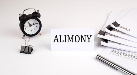 Card with text ALIMONY on a white background, near office supplies and alarm clock. Business concept.