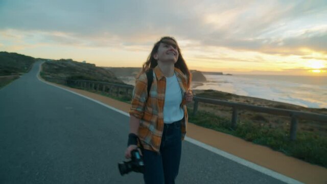 Female photographer walk on beautiful seaside road trail. Young woman on solo adventure road trip free and careless make photos of sun setting behind ocean landscape. Woman travel and create photos