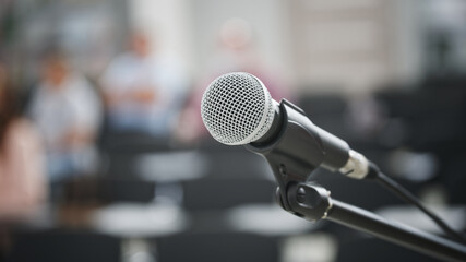 A microphone with a metal grill on a black stand against the background of people and the...