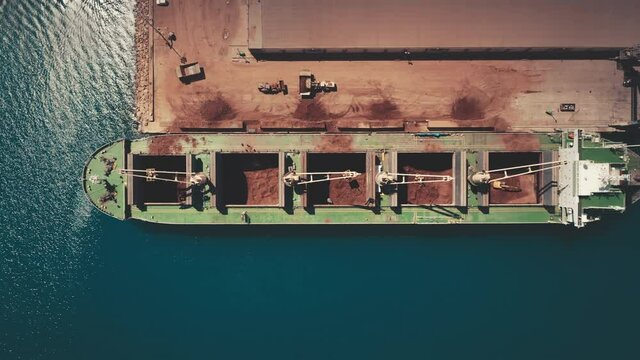 Aerial view, loading and unloading ship in roadstead at sea. Blue calm ocean water. Dry cargo ship with open holds is in port, zoom out. Cargo cranes are working at dock. Transportation goods.