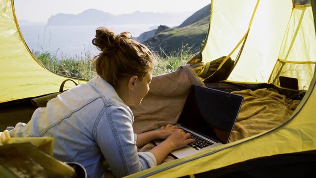 Young woman in jacket lies on blanket in yellow tent and types on grey laptop against rocky cliffs silhouettes in dense mist on sunny day close view