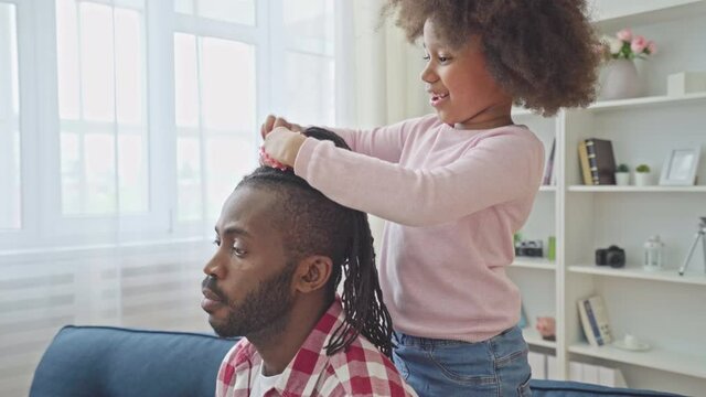 Cute daughter making hairstyle to patient father sitting on sofa, childhood