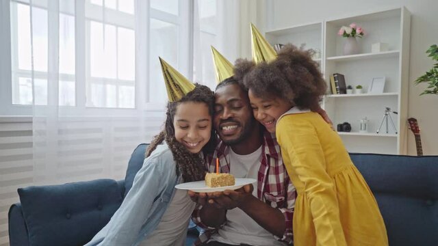 Happy black father blowing birthday cake candle, caring daughters hugging dad