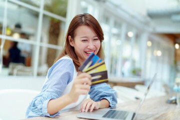 A beautiful Asian woman in a white shirt is sitting in front of a computer with a credit card in her hand to prepare for online shopping. Her face looks very happy in a bakery.