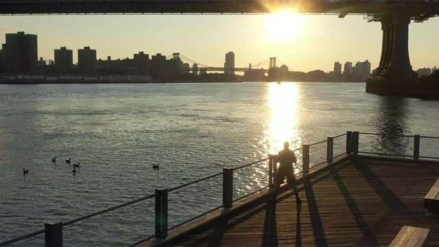 A  view of the East River at sunrise. The drone camera dolly in over a man, as it flies towards the Manhattan Bridge, the sun shines and reflects on the river in the early morning.