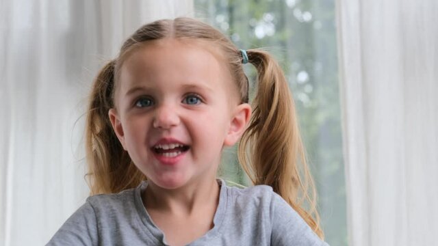Joyful naughty young girl with long pigtails screams and hits table with hands sitting at home against window close view