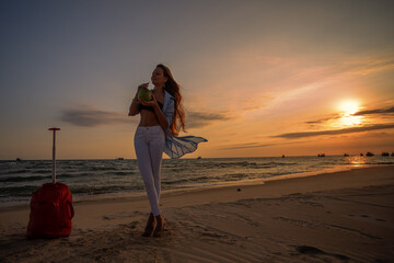 Young beautiful woman with large red suitcase  drink coconut on tropical beach