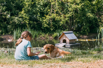 Friendship of animals and children, people. Caucasian girl sits on the bank of a pond, river and plays with a dog puppy, labrador retriever. Outdoor games, summer vacation