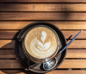 Cappuccino with latte art in glass cup on wooden table in outdoor cafe or coffeehouse. Sunny morning. Top view.