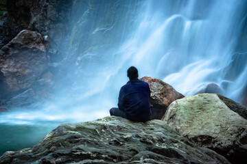 Naklejka na ściany i meble man sitting at rock in front of waterfall white water stream falling from mountains long exposure