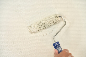  A man's hand uses a paint roller to apply light paint to the glass wallpaper on the wall. Copy...