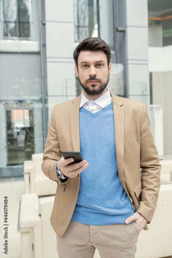 Aufkleber portrait of young bearded businessman using mobile phone and looking at camera while standing at off