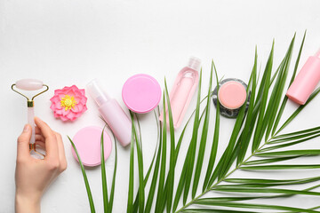 Female hand with facial massage tool, cosmetic products and palm leaf on light background
