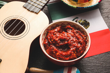 Bowl of tasty salsa sauce with symbols of Mexico on dark background