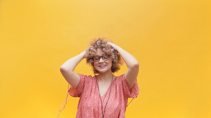 Attractive girl wearing nerdy spectacles with both hands on her head and smiling. Girl in a joyful mood and wearing earphones listening to her favorite music. Isolated in a bright yellow background.