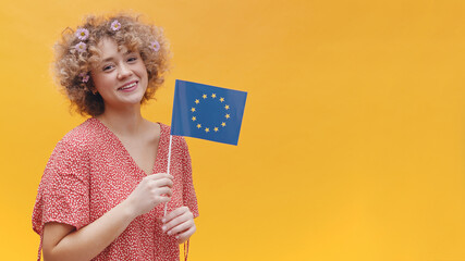 Girl holding a small European Union flag in her hand. The girl is isolated over a bright yellow background with EU Flag. Symbol of Europe, EU association.