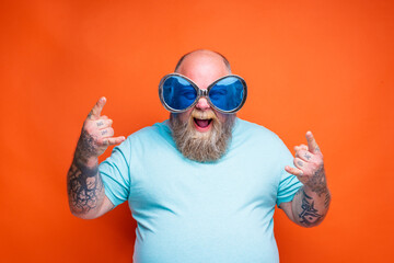Fat happy man with beard, tattoos and sunglasses makes the gesture of the horns
