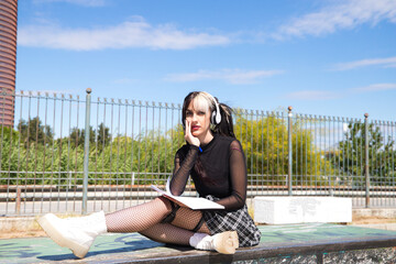 young and beautiful university girl with a punk tendency. she is sitting in the park. she has a notebook and a pen in her hands and is doing her homework to take to class.