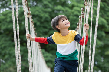 A boy wearing bright colored clothes walking alone on rope bridge at extreme sport in adventure park.