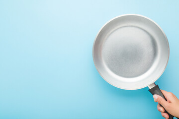 Young adult woman hand holding new aluminium frying pan on light blue table background. Pastel color. Closeup. Cooking concept. Empty place for text. Top down view.