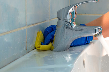 Cleaning of inter tile joints in bathroom from dirt and black poisonous mold in hard to reach place behind tap. Hand in blue glove uses yellow rag and detergent to wash dirt behind faucet in bathroom.