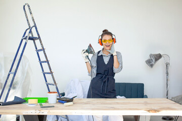 Young dark-haired woman in glasses and headphones smiling running power tools in workshop preparing wooden table surface to painting