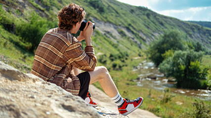 A young man with curly hair taking shots using camera in the nature, sitting on a rock, greenery and river nearby © frimufilms