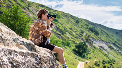 A young man with curly hair taking shots using camera in the nature, sitting on a rocky hill's slope