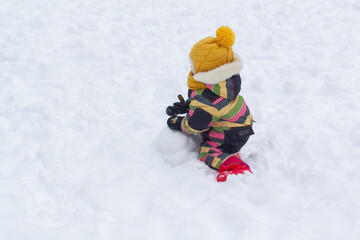 Fototapeta na wymiar A child girl in a bright winter jumpsuit sculpts a large lump of snow in the winter on the playground