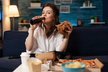 Portrait of woman looking into camera during fastfood lunch meal order relaxing on sofa late at night in living room. Caucasian female enjoying tasty burger, takeaway food home delivered