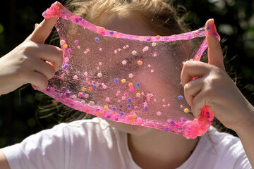 little girl shows transparent pink slime with decorations, looks through its texture