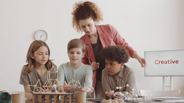 Tracking right of curly Caucasian woman standing behind diverse elementary school students who sitting at table, helping them assemble craft miniature model of building
