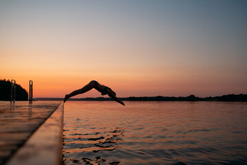 Obraz premium Woman jumping into the lake from wooden pier. Having fun on summer day on the vacations. Young girl diving in to the water from dock. Doing sports at dawn. Silhouette of a swimmer against sunset sky.