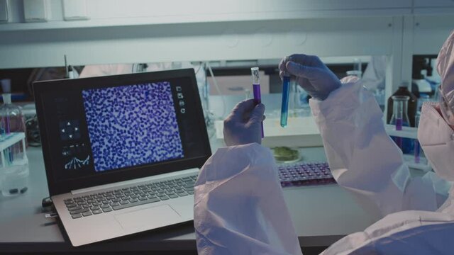 Slowmo PAN of female scientist in disposable coveralls, goggles and face mask looking at solution in test tubes while doing research in lab
