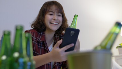 Young happy attractive asia female enjoy smile laugh relax night party event online celebration festive with friends at home sofa couch living room drink beer with glass and bottle online videocall.
