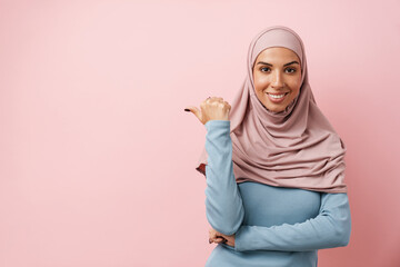 A smiling muslim woman wearing pink hijab pointing to the copyspace