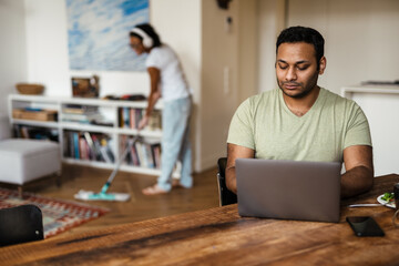 Middle eastern man working with laptop while his girlfriend cleaning floor