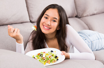 Obraz na płótnie Canvas Positive young female eating salad witn fork lying at home